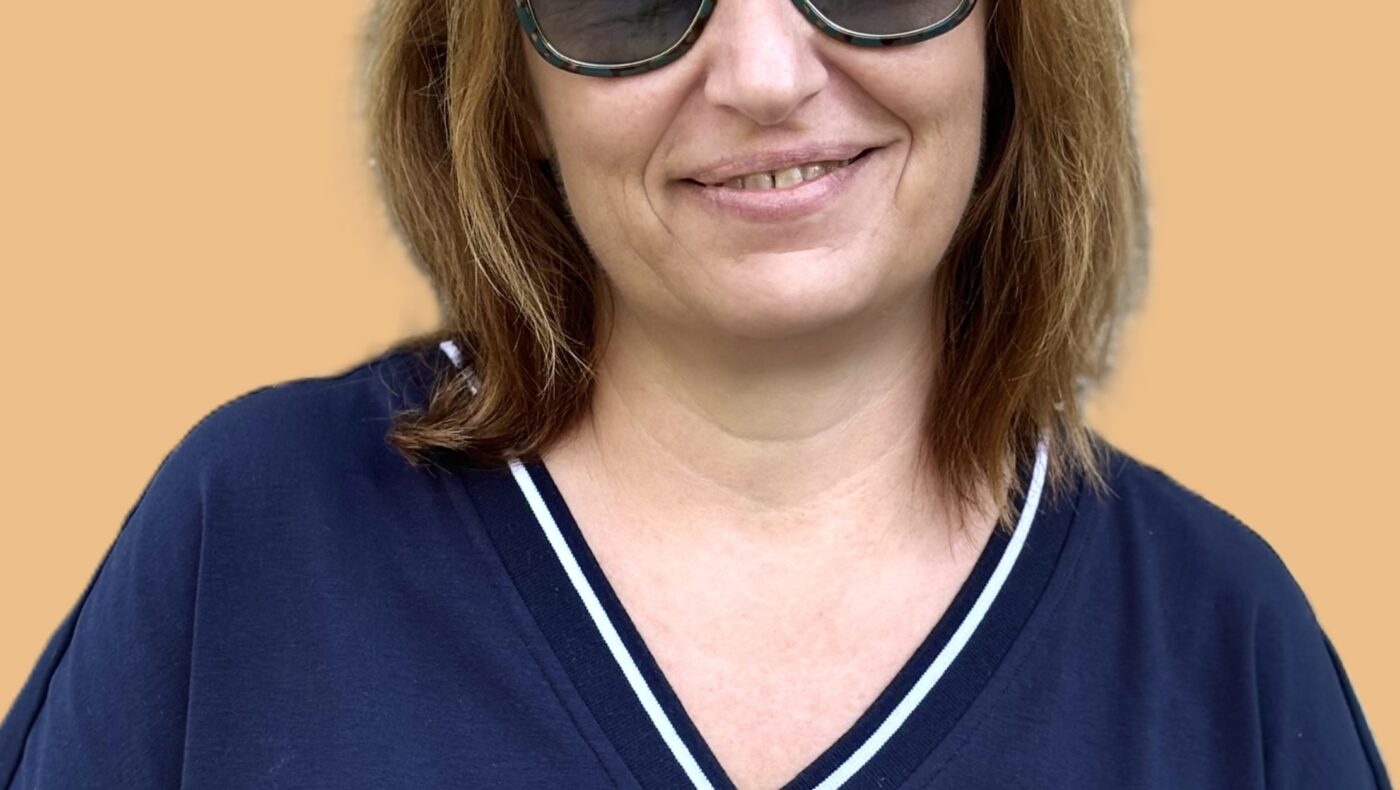 Woman with sunglasses and brown hair wearing a navy top, standing and smiling against light orange background.