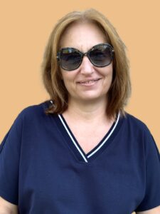 Woman with sunglasses and brown hair wearing a navy top, standing and smiling against light orange background.