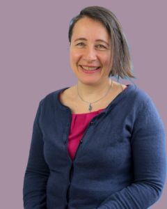 Female teacher smiling while wearing a navy cardigan and pink top against a light purple background.