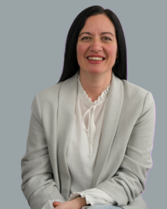 Woman with long dark hair wearing a light gray blazer and white blouse against a purple background.