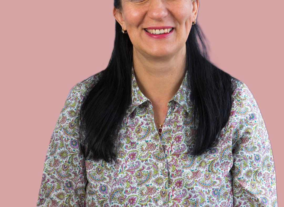 Woman with long dark hair wearing a patterned shirt, smiling against a pink background.