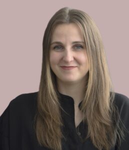 A woman with long hair wearing a black shirt, smiling at the camera against a neutral background.